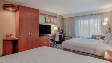 A hotel room with two beds, a desk with a TV, a minibar, and a large window with curtains.