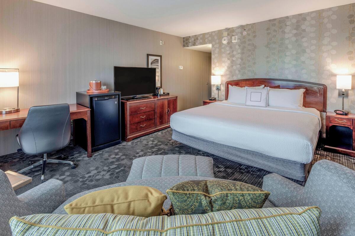 A hotel room with a large bed, a desk with a television, a chair, and a sofa with cushions.