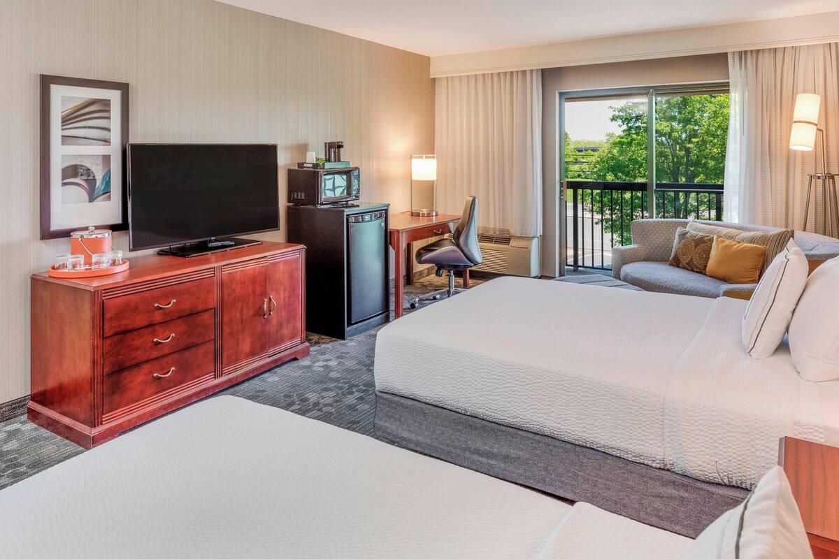 A hotel room with a large bed, a desk with a television, a dresser, and a balcony with a view of trees.