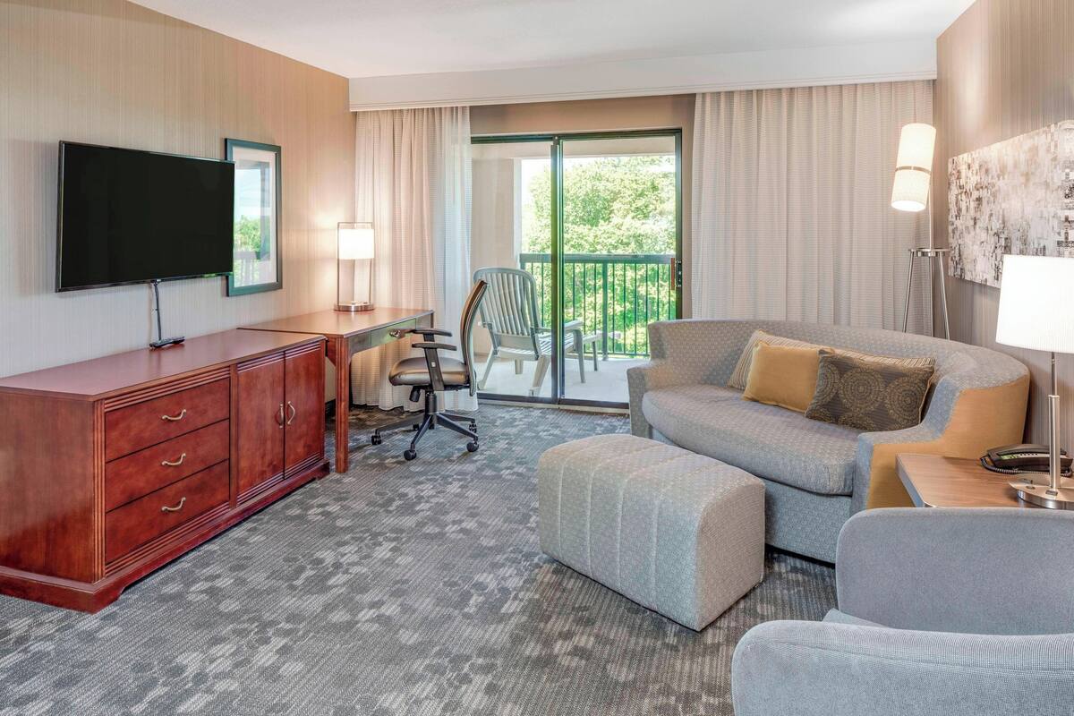 A hotel room with a flat-screen TV, a desk with a chair, a sofa, and a balcony with a view of greenery.