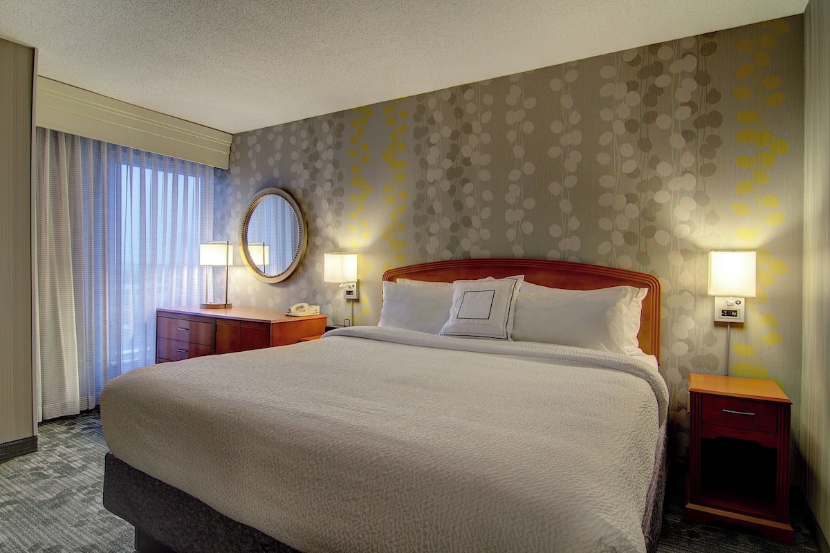A hotel room with a large bed, two bedside tables with lamps, a round mirror, and a window with curtains.