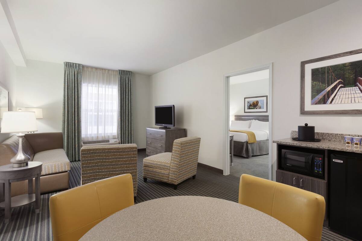 A hotel room with a bed, a desk, a chair, a dining table, a TV, a microwave, and a kitchen area.
