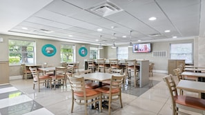 A spacious dining area with multiple tables and chairs, large windows, and a television screen on the wall.
