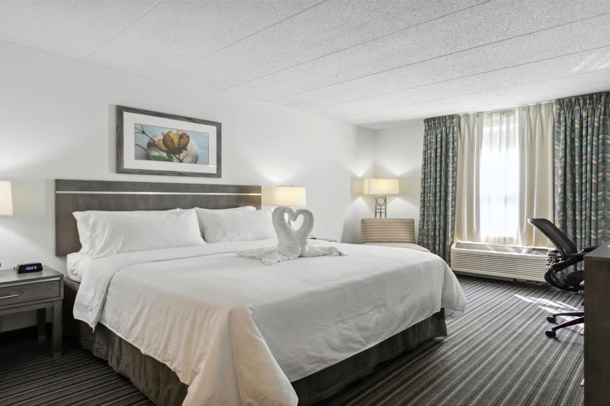 A hotel room with a large bed, two bedside tables with lamps, a desk with a chair, a framed picture on the wall, and a window with curtains.