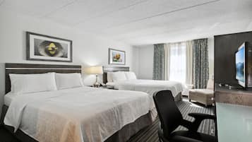 Habitación de hotel con dos camas, un escritorio con computadora, una silla, televisor y obras de arte en las paredes.