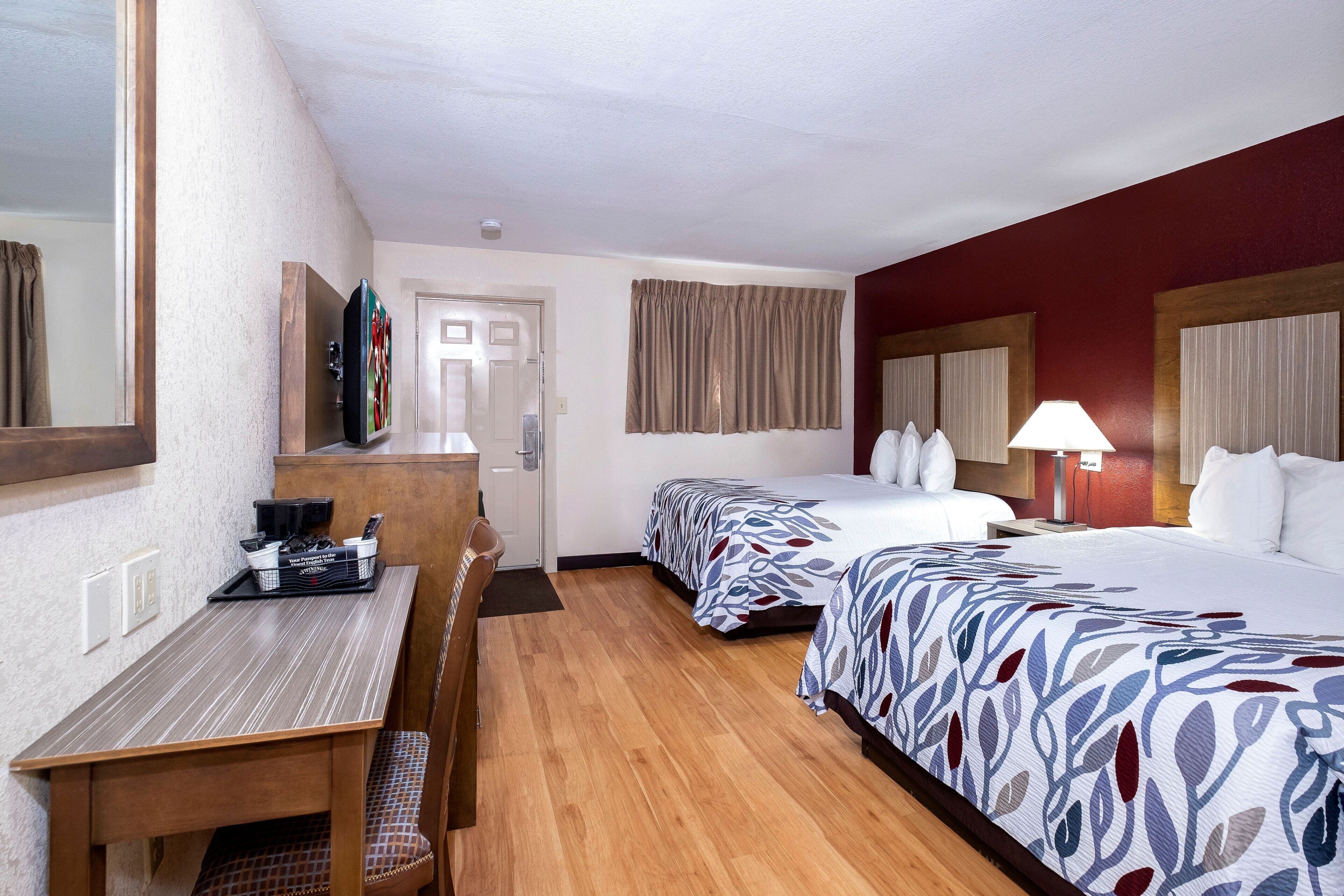 Habitación de hotel con escritorio, dos camas, un espejo, una puerta y una ventana con cortinas.