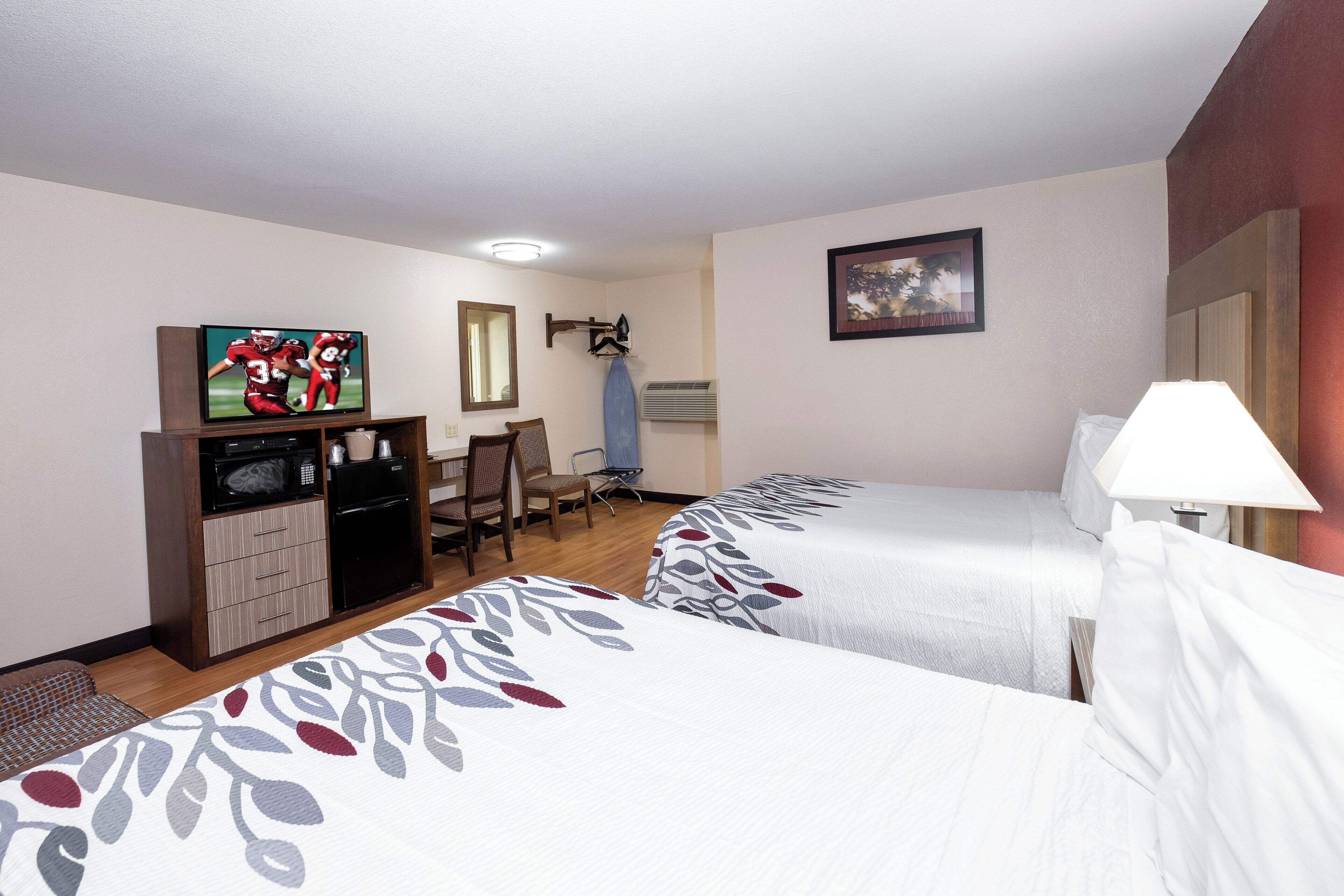 Habitación de hotel con dos camas, un escritorio con televisor, una silla, una lámpara de escritorio y un cuadro en la pared.