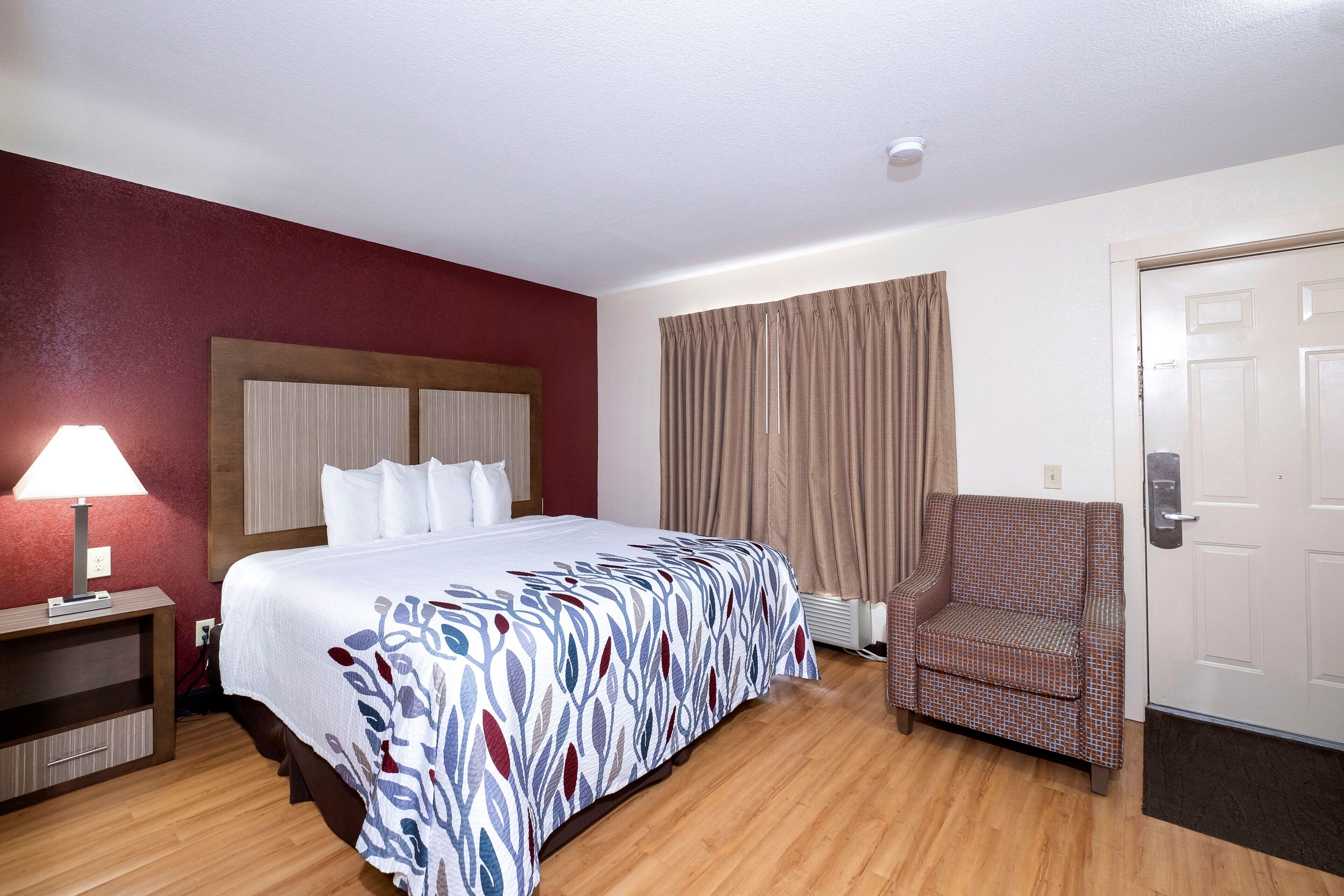 A hotel room with a bed, a chair, a lamp, and a door.