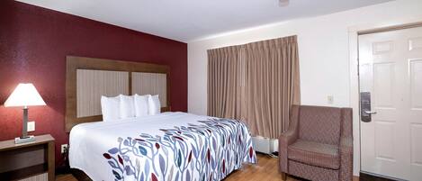 A hotel room with a bed, a chair, a lamp, and a door.