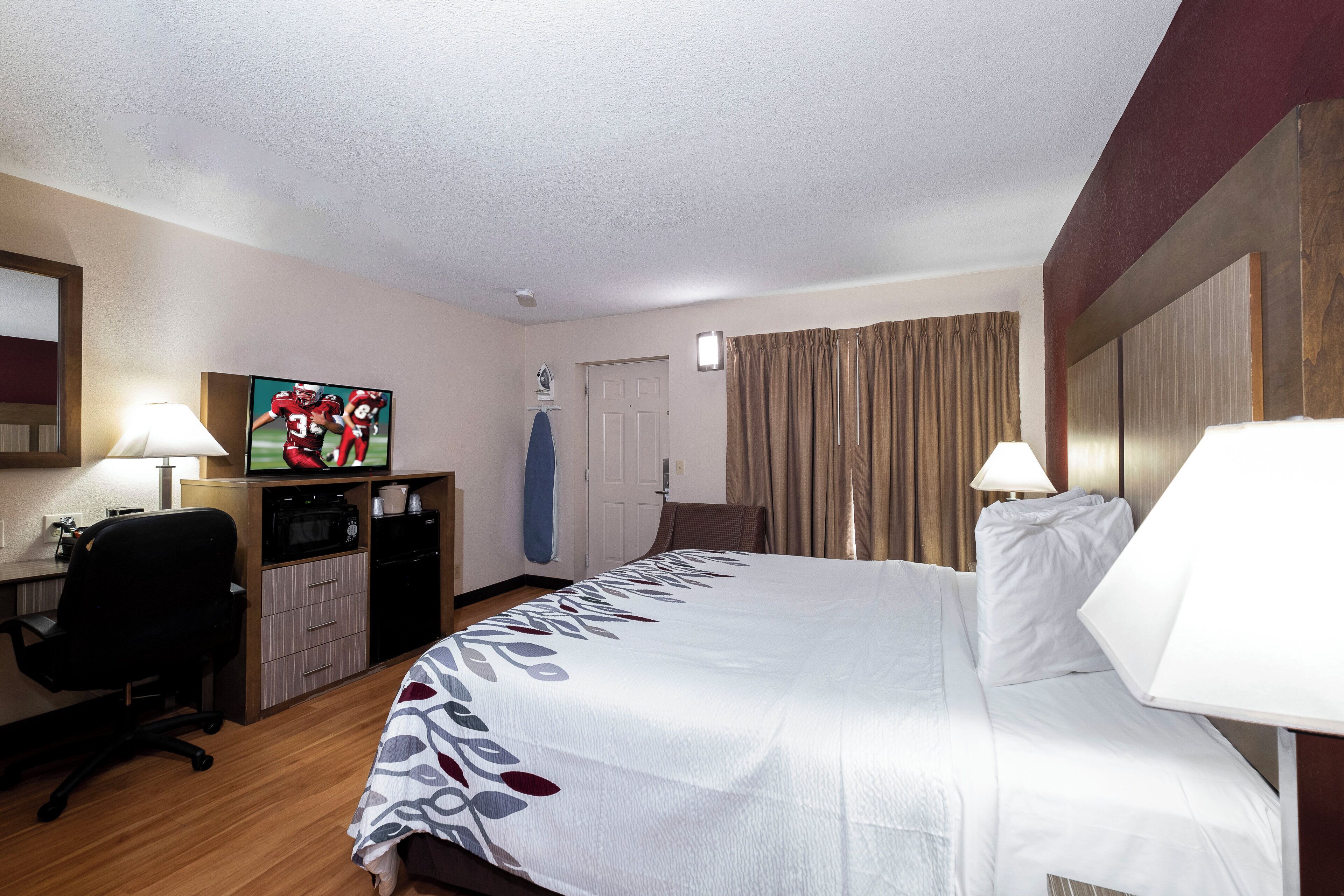 A hotel room with a bed, a desk with a television, a chair, a microwave, and a door.