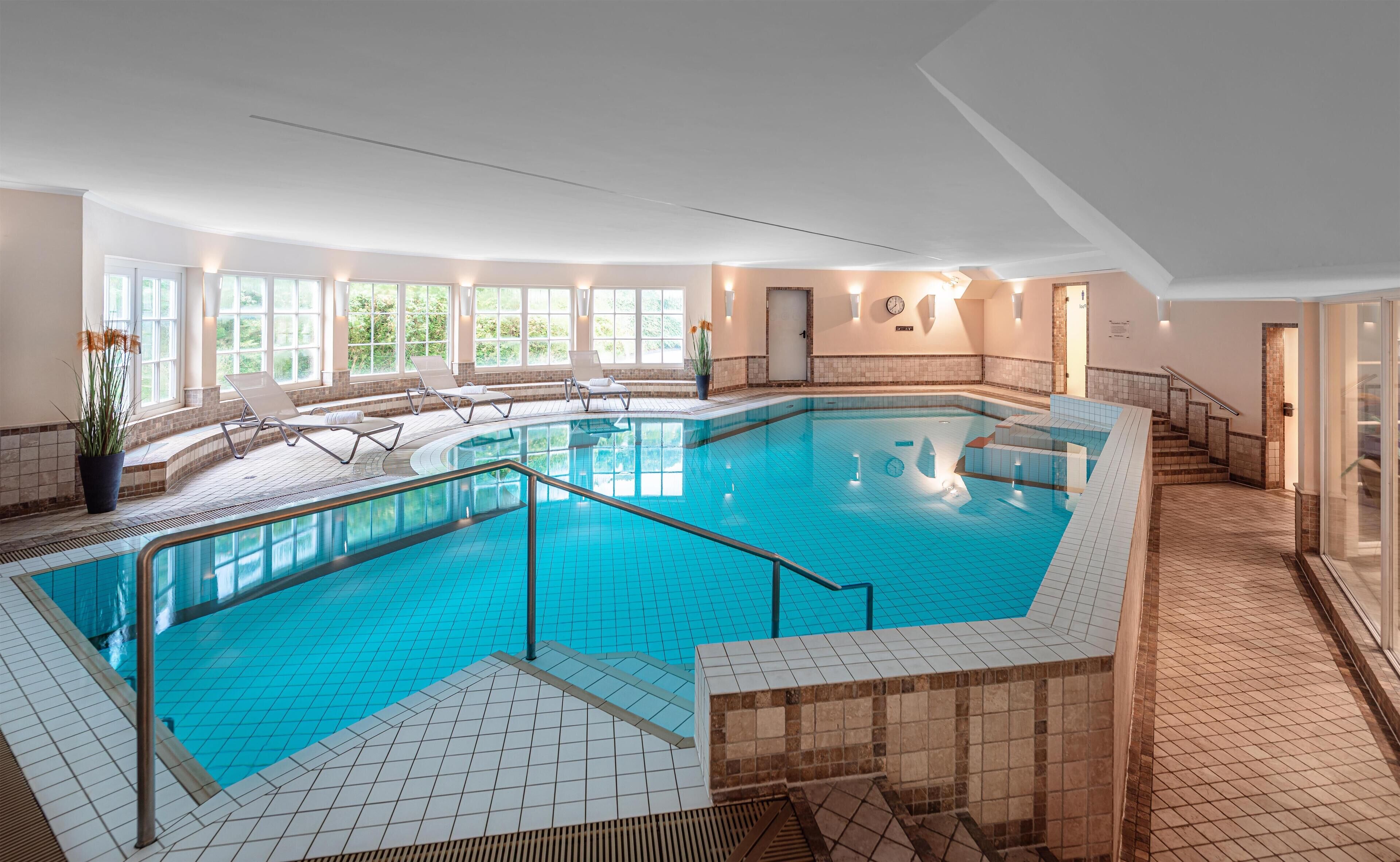 A large indoor swimming pool with a tiled floor, sun loungers, and a staircase leading to a door.