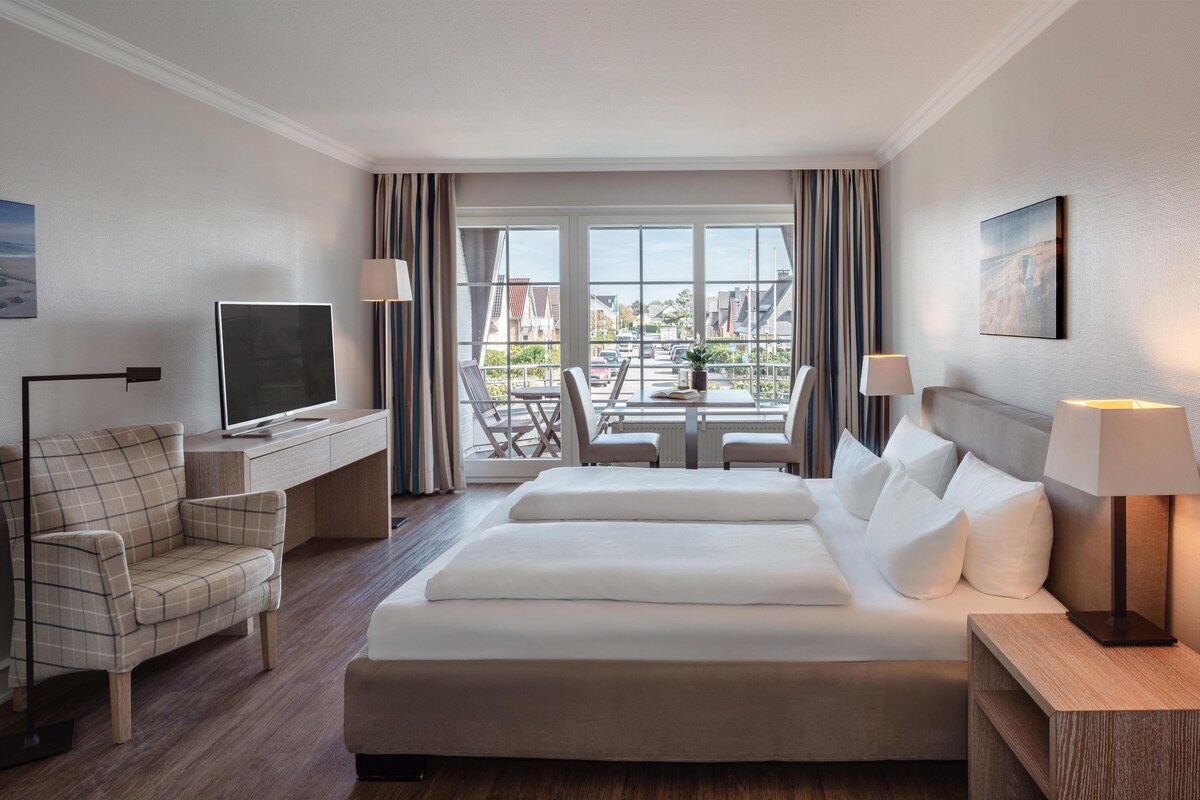 A hotel room with a bed, a desk with a TV, a chair, a lamp, and a view of the outside.