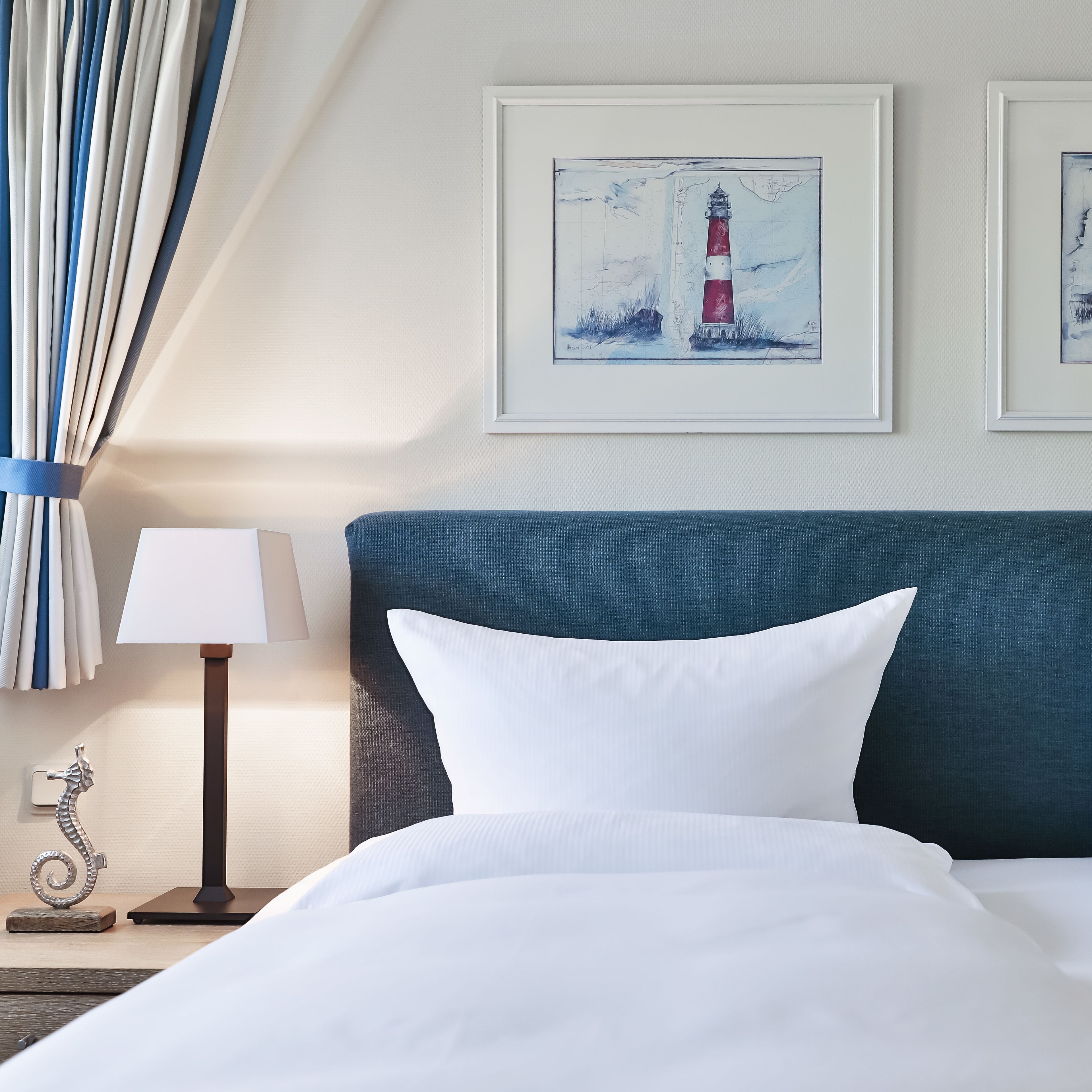 A neatly made bed with white bedding, a blue headboard, and two framed artworks depicting lighthouses on the wall.
