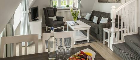 A modern living room with a dining area, featuring a wooden table with a fruit platter, a white coffee table, and a sofa set.