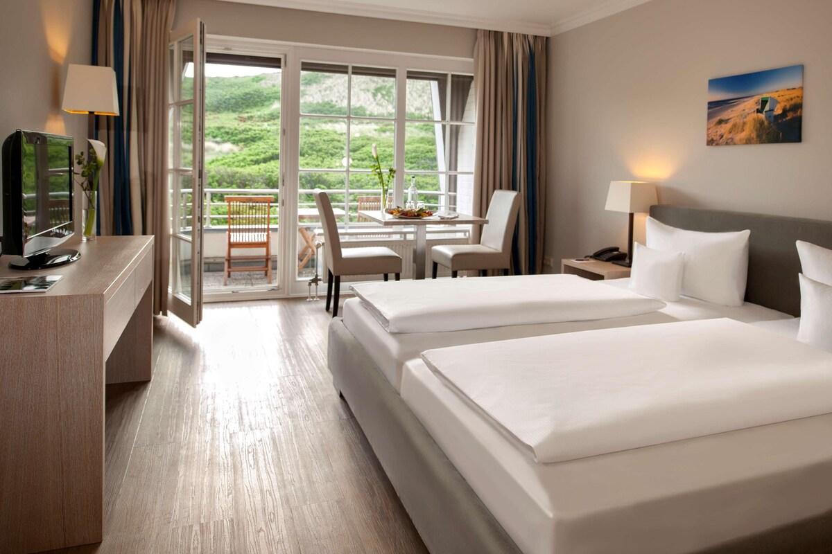 A hotel room with two beds, a desk with a computer, a chair, a table with a vase, and a balcony with a view of greenery.