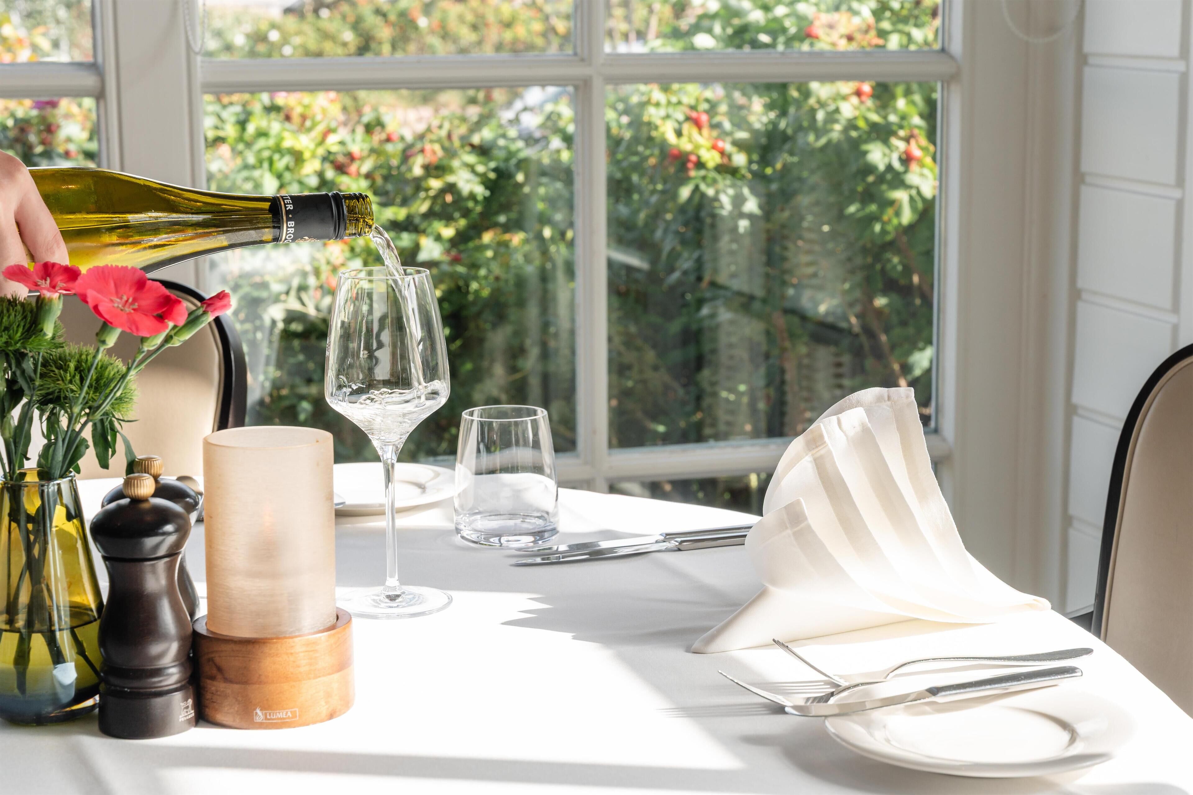 A table set for dining with a bottle of wine being poured, a candle, and a vase with flowers.