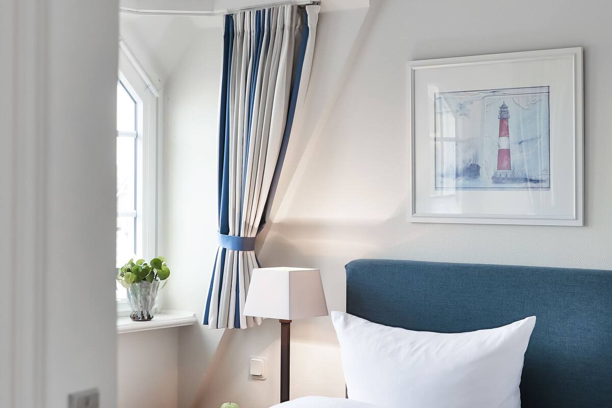 Et hotelværelse med seng, et vindue med gardiner, en vægmonteret lampe og et indrammet billede.