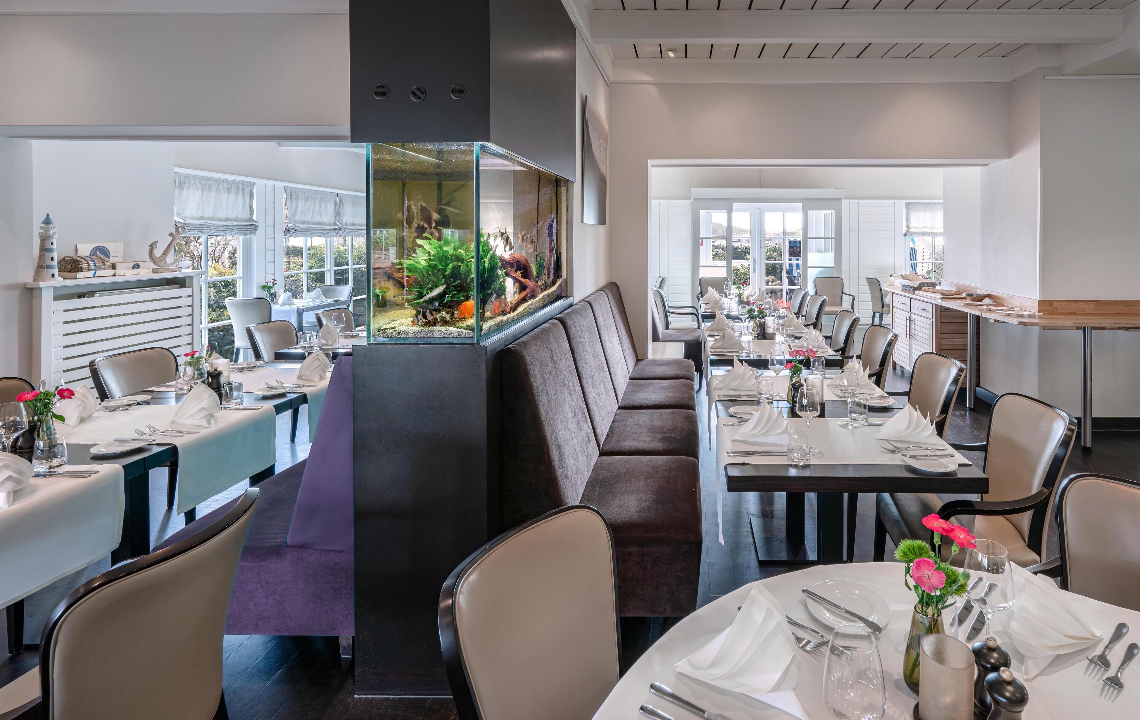 A modern dining area with multiple tables set for service, a large aquarium, and a view of the outdoors.