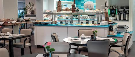 A modern hotel dining area with round tables, chairs, a buffet counter, and a kitchen in the background.