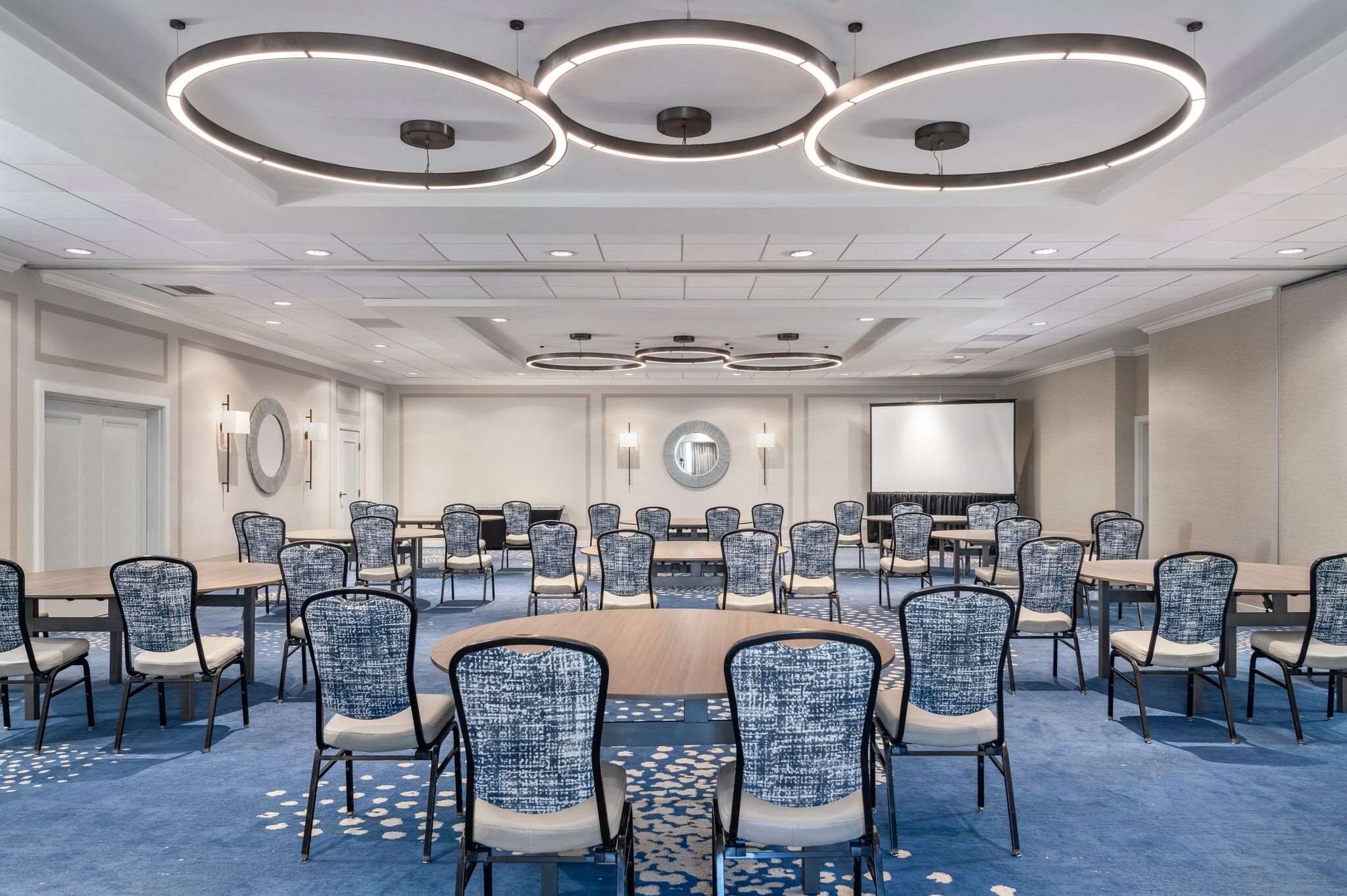 A conference room with round tables and chairs, a large screen, and circular ceiling lights.