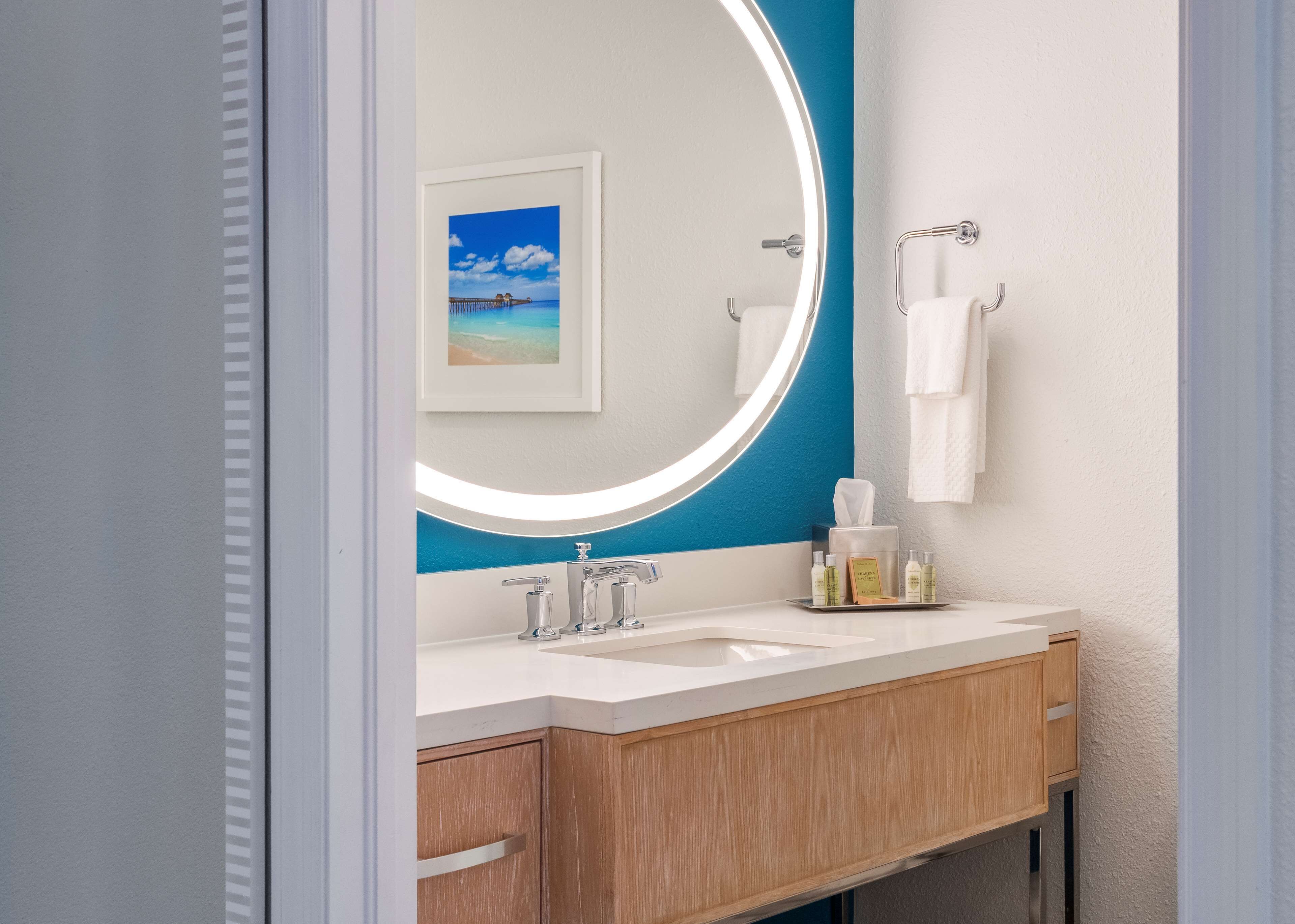 A bathroom with a white sink, two faucets, a mirror with a blue accent, a framed picture of a beach, and a towel rack.