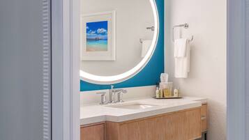 A bathroom with a white sink, two faucets, a mirror with a blue accent, a framed picture of a beach, and a towel rack.