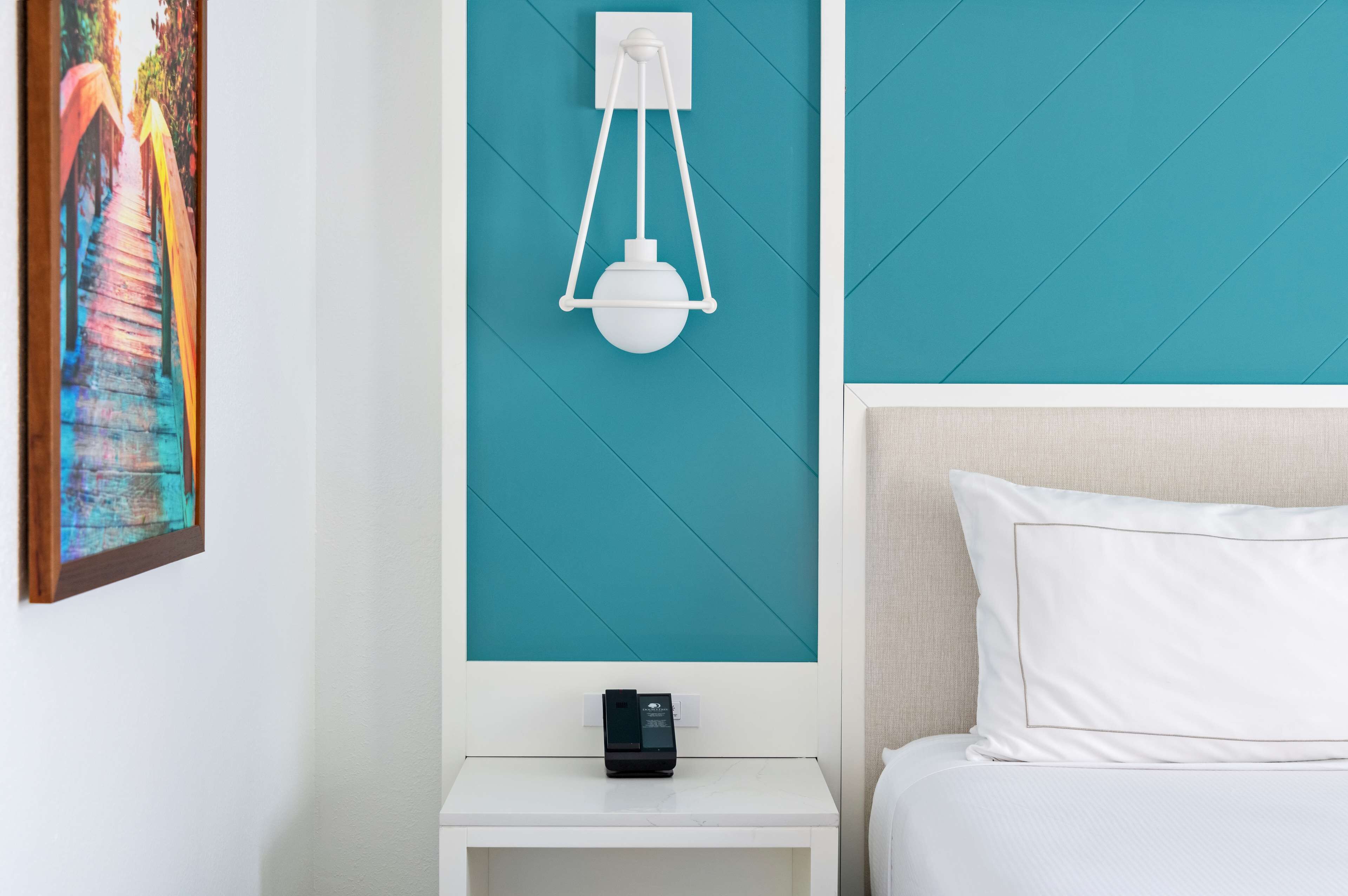 A hotel room with teal walls, a white bedside table, a white bed with a white pillow, and a wall-mounted light fixture.