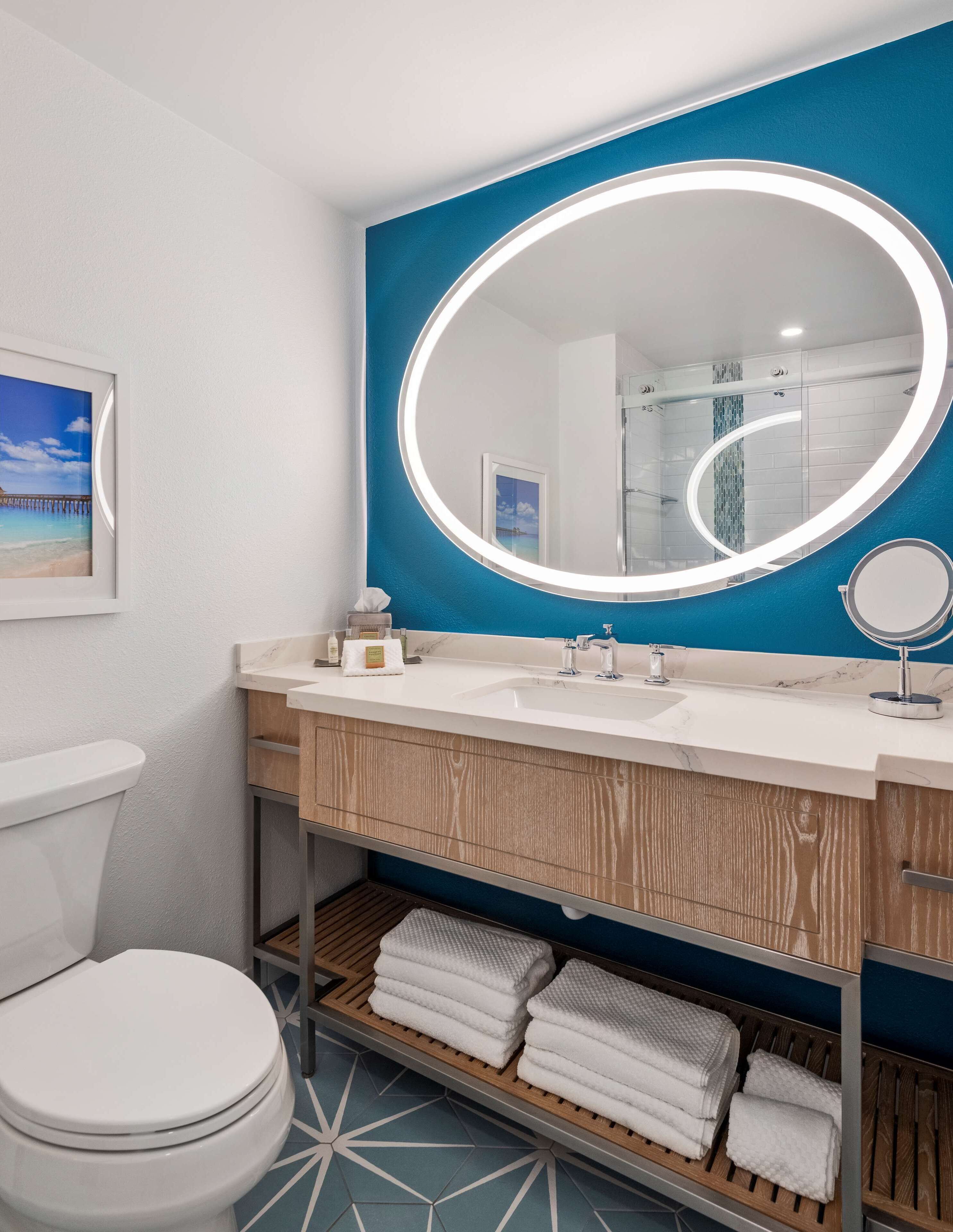 Uma casa de banho moderna com uma parede azul de destaque, um espelho redondo com iluminação em halo, uma sanita branca e uma prateleira com toalhas dobradas.