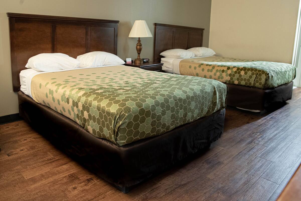 Habitación de hotel con dos camas, cabeceros de madera, una mesita de noche con lámpara y suelo de madera.