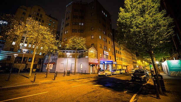 A nighttime street view with a building featuring multiple floors and a variety of shops on the ground floor. - Travelodge Belfast Central (Belfast)