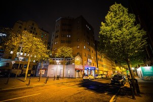 Une vue nocturne d’une rue, avec un immeuble à plusieurs étages et divers commerces au rez-de-chaussée.
