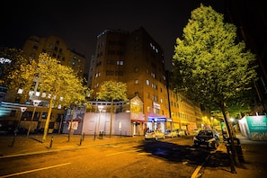 Vista nocturna de una calle con un edificio de varios pisos y diversas tiendas en la planta baja.