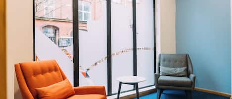 A room with a blue carpet, two chairs, a small table, and large windows with a view of buildings outside.