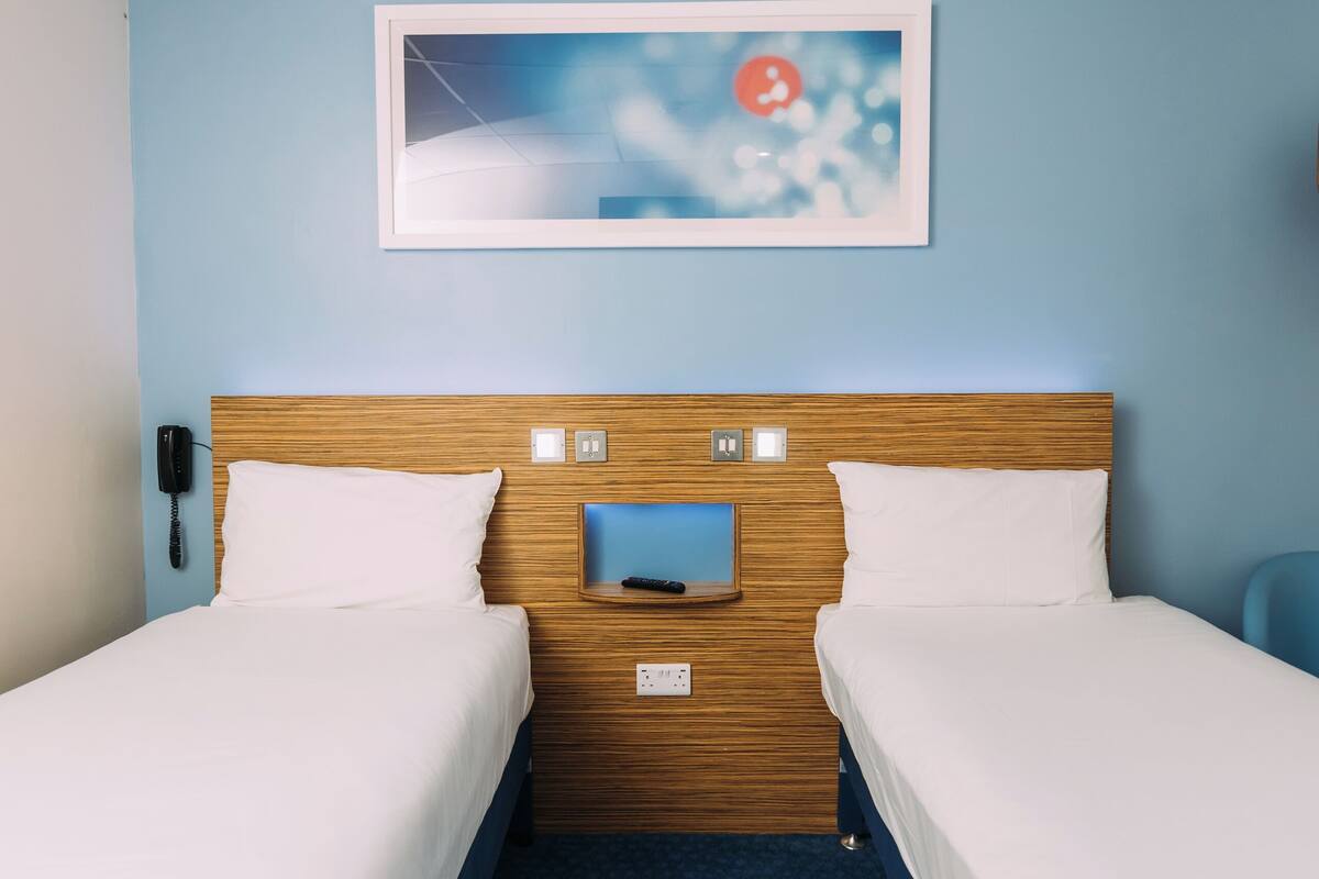 Une chambre d’hôtel avec deux lits simples, une tête de lit en bois, une petite table de chevet, un téléphone et un tableau encadré au mur.