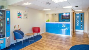 A reception area with a blue counter, a red chair, a blue chair, a vending machine, and a sign for rooms. - Travelodge Burton M6 Northbound (Carnforth)