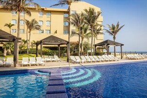 Piscina com espreguiçadeiras e palmeiras, cercada por um edifício com várias janelas.
