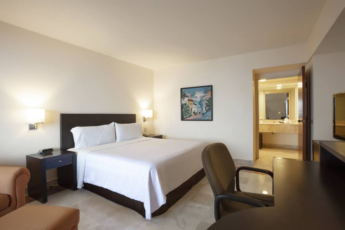 Habitació d'hotel amb llit, taules de nit, una cadira, escriptori i bany amb mirall.