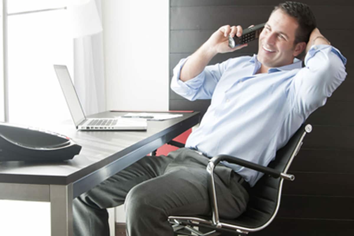 A man sitting in a modern office chair at a desk with a laptop, a smartphone, and a briefcase.