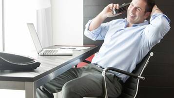 En man sitter på en modern kontorsstol vid ett skrivbord med en bärbar dator, en smartphone och en portfölj.
