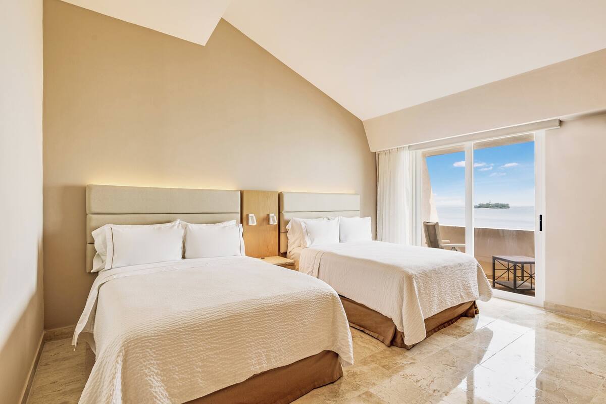 Habitació d'hotel amb dos llits, una finestra gran amb vistes i un balcó amb taula i cadires.