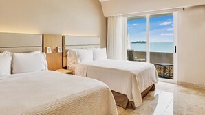Habitación de hotel con dos camas, una mesita de noche, una mesa y vista al mar.