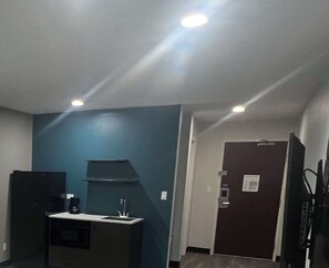 A small, dimly lit room with a kitchenette area, a dark door, and a ceiling light. - Studio 6 Oklahoma City - Moore/Norman (Oklahoma City)