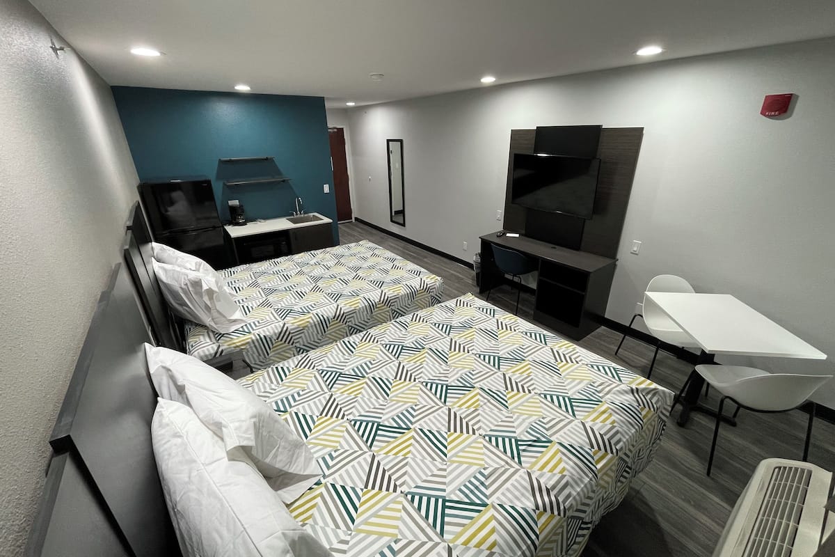 Ein Hotelzimmer mit zwei Betten, einem Schreibtisch, einem Stuhl, einem Fernseher und einer kleinen Küchenzeile.