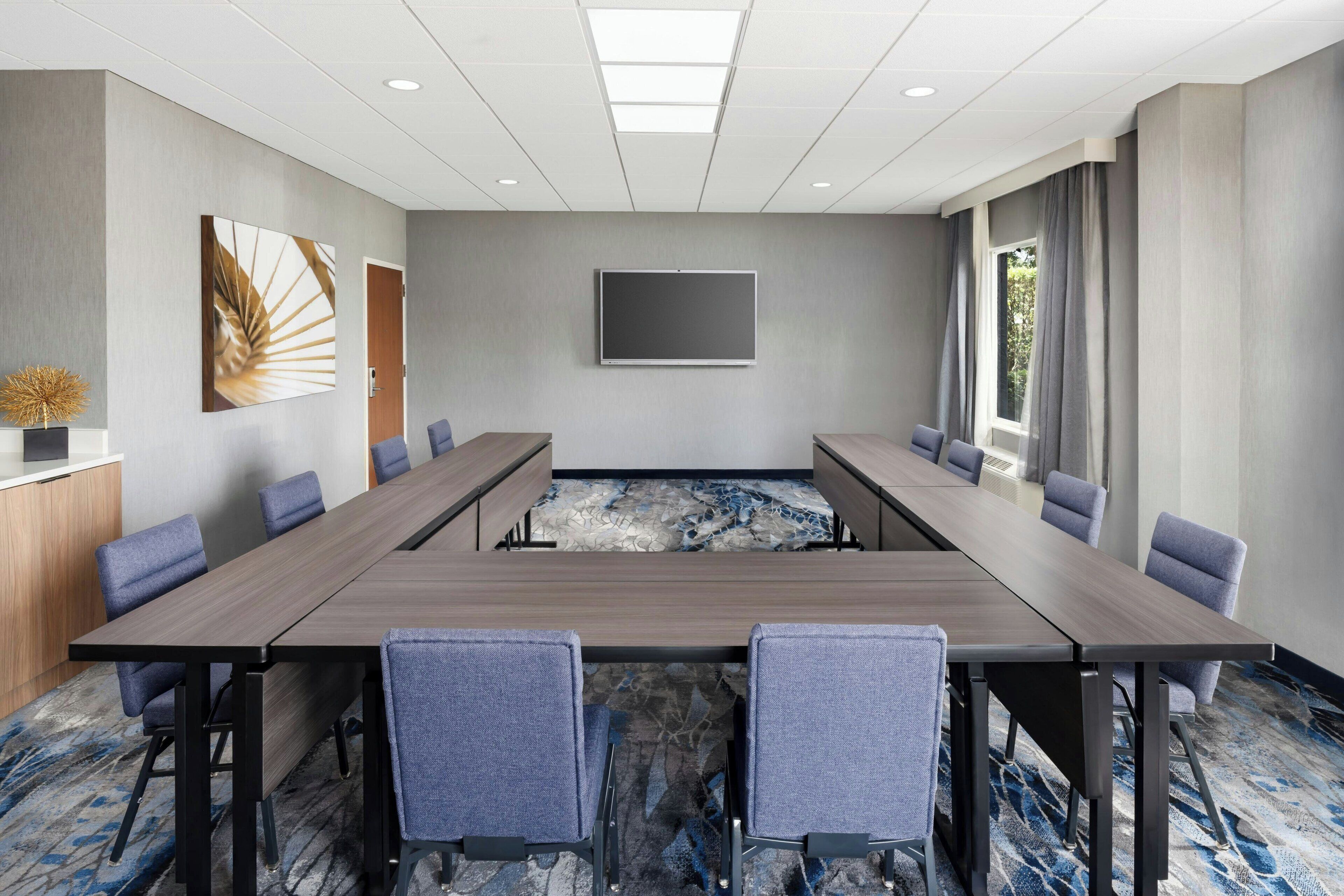 A conference room with a long table, multiple chairs, a flat-screen TV, and a window with curtains.