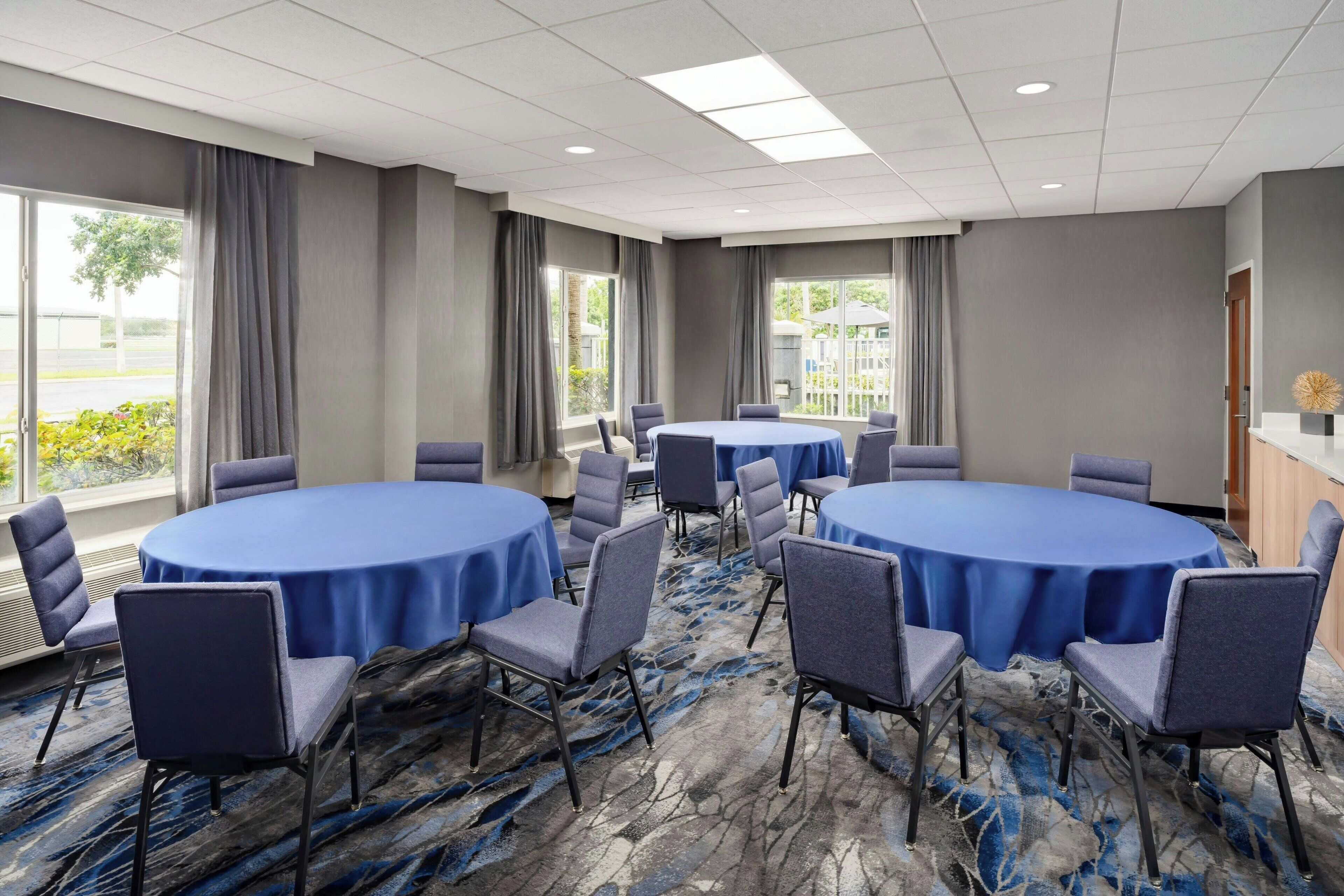 A conference room with round tables and chairs, large windows with curtains, and a carpeted floor.