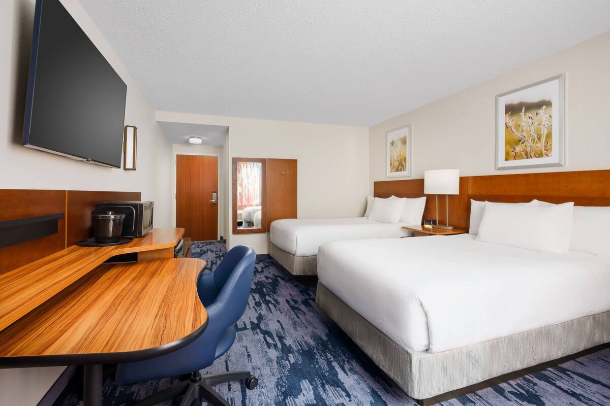 Habitación de hotel con un escritorio de madera, dos camas, un televisor y obras de arte en las paredes.