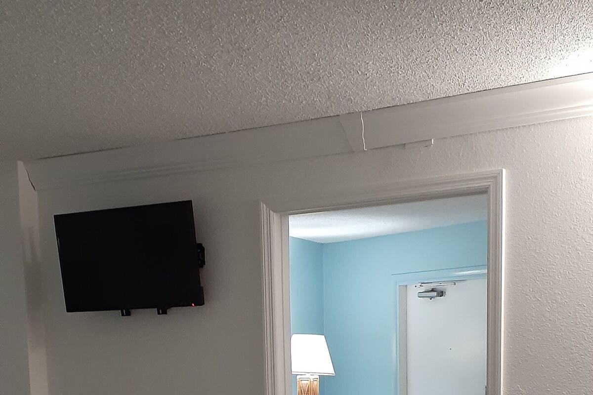 A bedroom with a bed, a TV mounted on the wall, a chair, and a door leading to another room.
