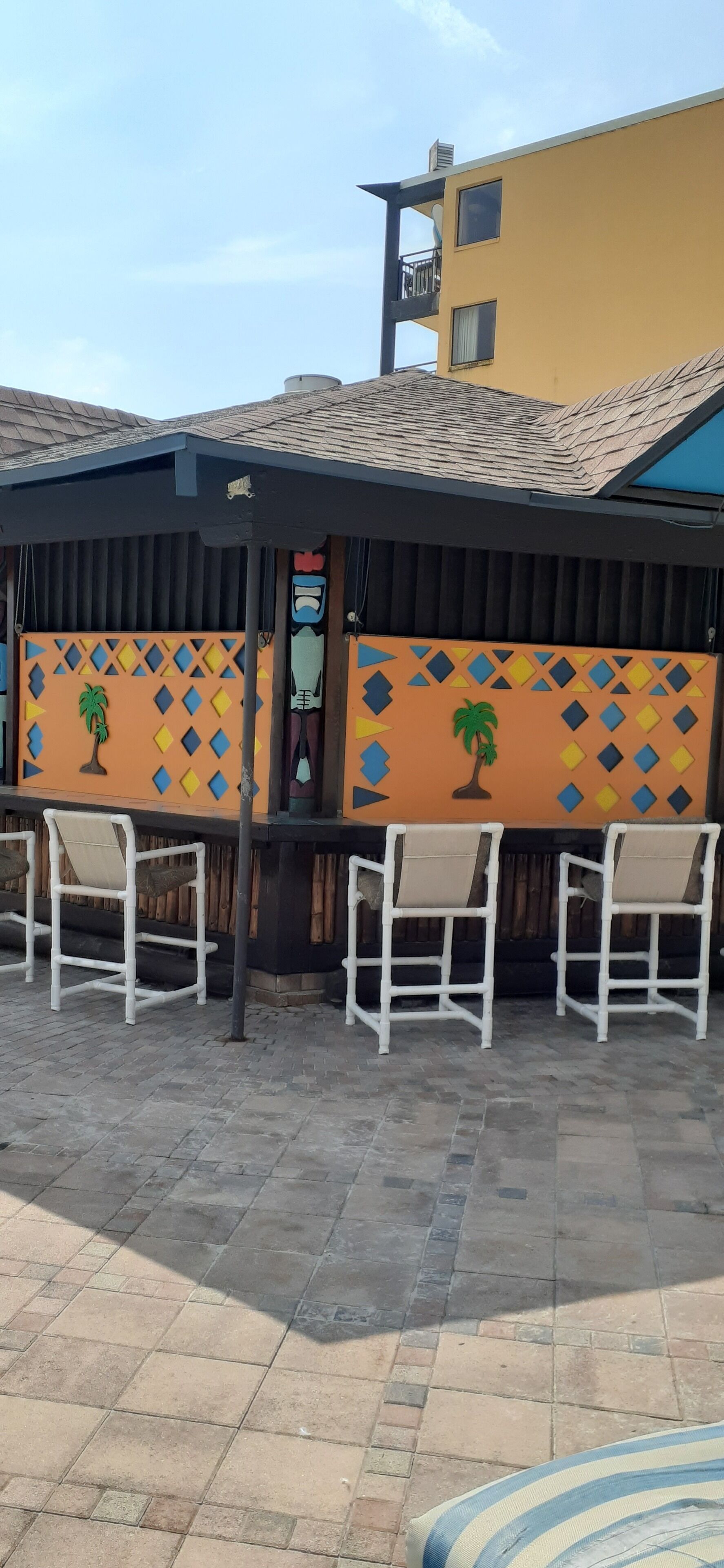 Un espacio al aire libre con techo, con sillas blancas y una pared decorativa con palmeras y diseños geométricos.
