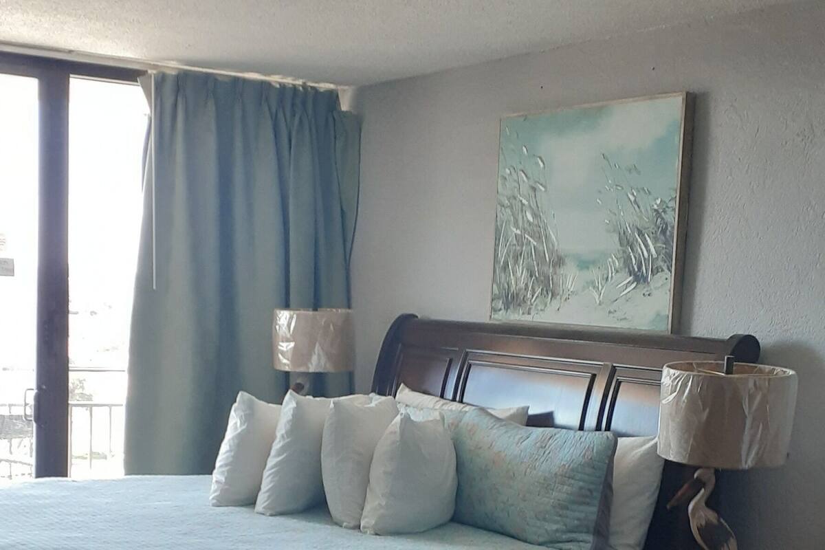 A bedroom with a bed, a nightstand, a lamp, a ceiling fan, and a window with curtains.