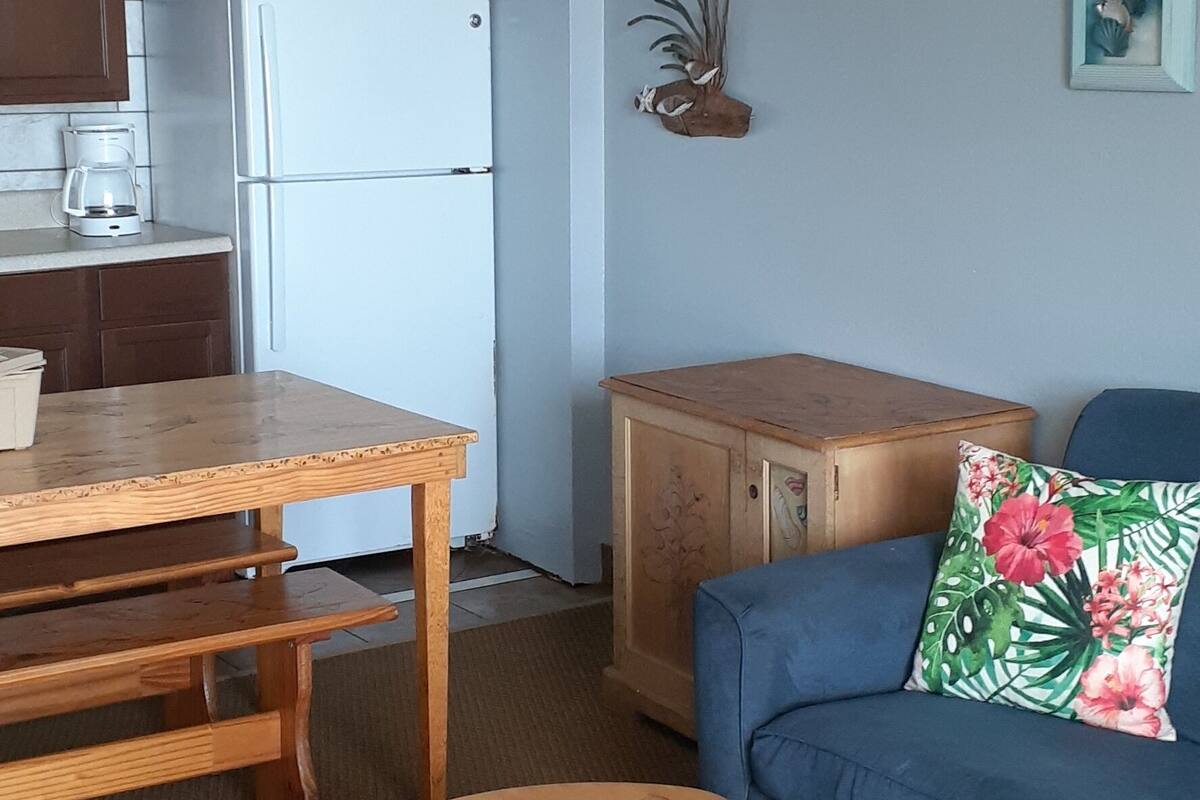 A compact living space with a kitchenette, a wooden dining table, a blue sofa with a floral pillow, and a small wooden bench.